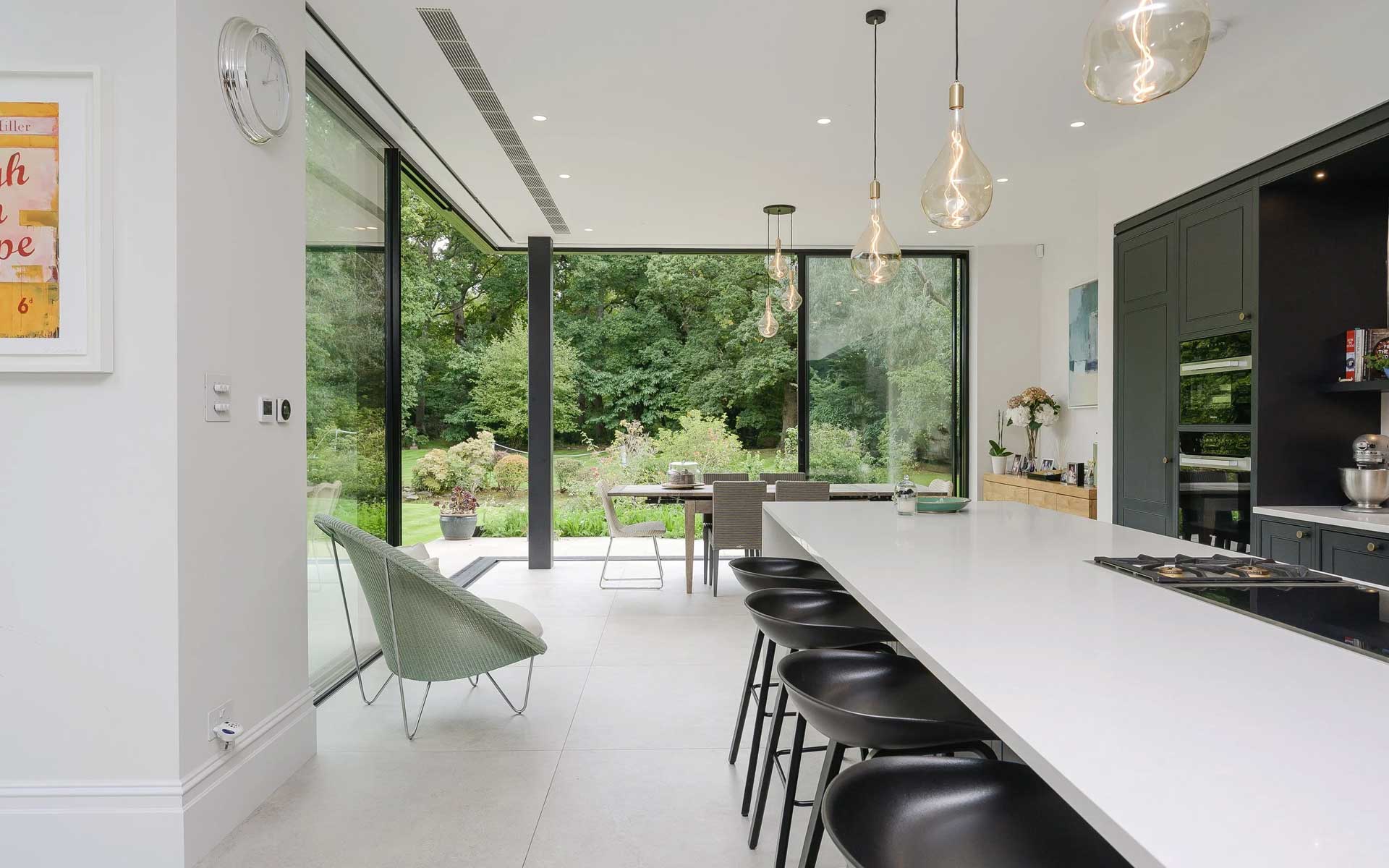 Long kitchen island with black stools facing sliding glass doors opened onto a garden terrace and mature trees
